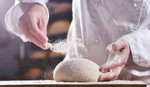 Homem com traje de badeiro, espalhando farinha de trigo sobe a massa de pão que está sobre uma mesa